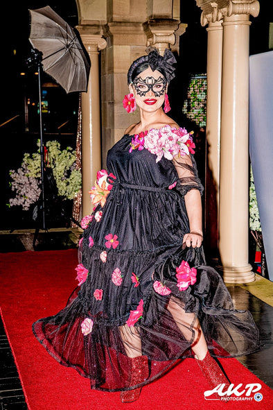 Picture of model Andrea at Ravishing Fashionistas Spring Fashion Show. She is wearing blacking netting dress with spring flowers across her shoulders and skirt. Her earrings are handmade clip on flowers and a black diamond mask and headband. .